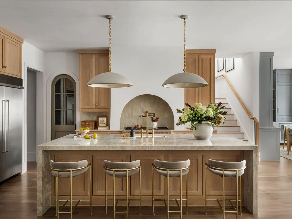 Modern kitchen with a large marble island, four bar stools, pendant lights, a flower vase, and wooden cabinets.