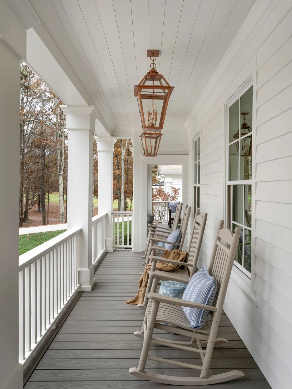 A porch with white railings, columns, and walls, decorated with hanging lantern-style lights, and four rocking chairs with pillows and a blanket facing the yard with trees.