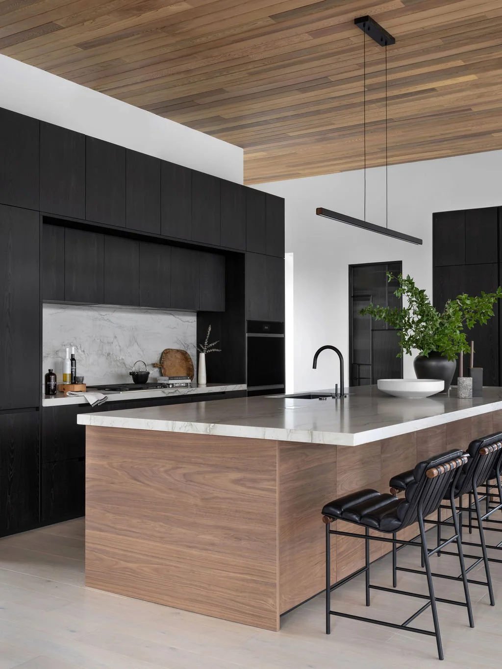 Modern kitchen with black cabinetry, white marble backsplash, wooden ceiling, and a kitchen island with a marble top. The island has black barstools and a large green plant in a black vase.