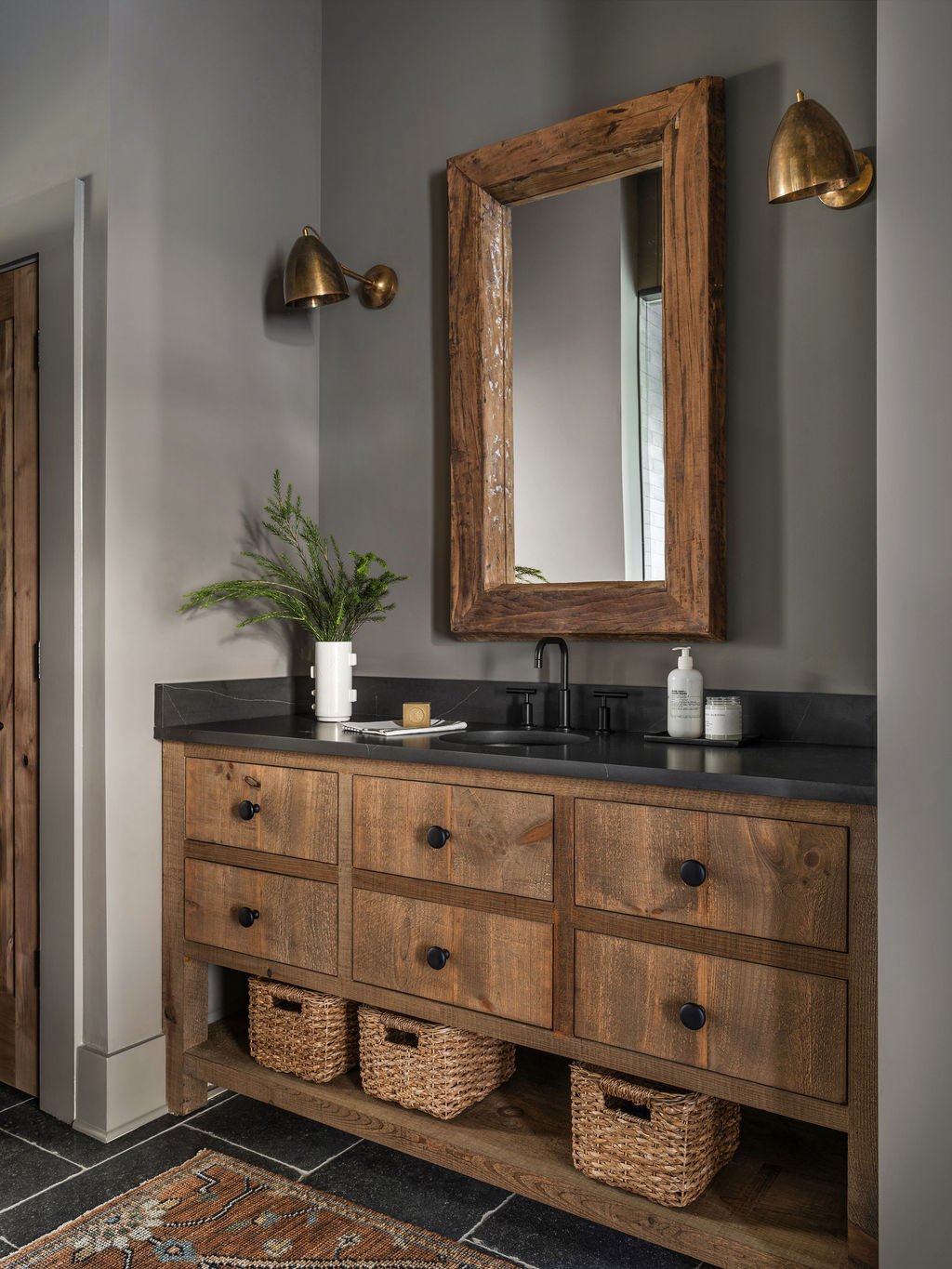 Rustic bathroom with a wooden vanity, black countertop, and a large wooden framed mirror. Two brass wall sconces are mounted on gray walls. A potted green plant sits on the vanity along with toiletries, and woven baskets are stored on the lower shelf