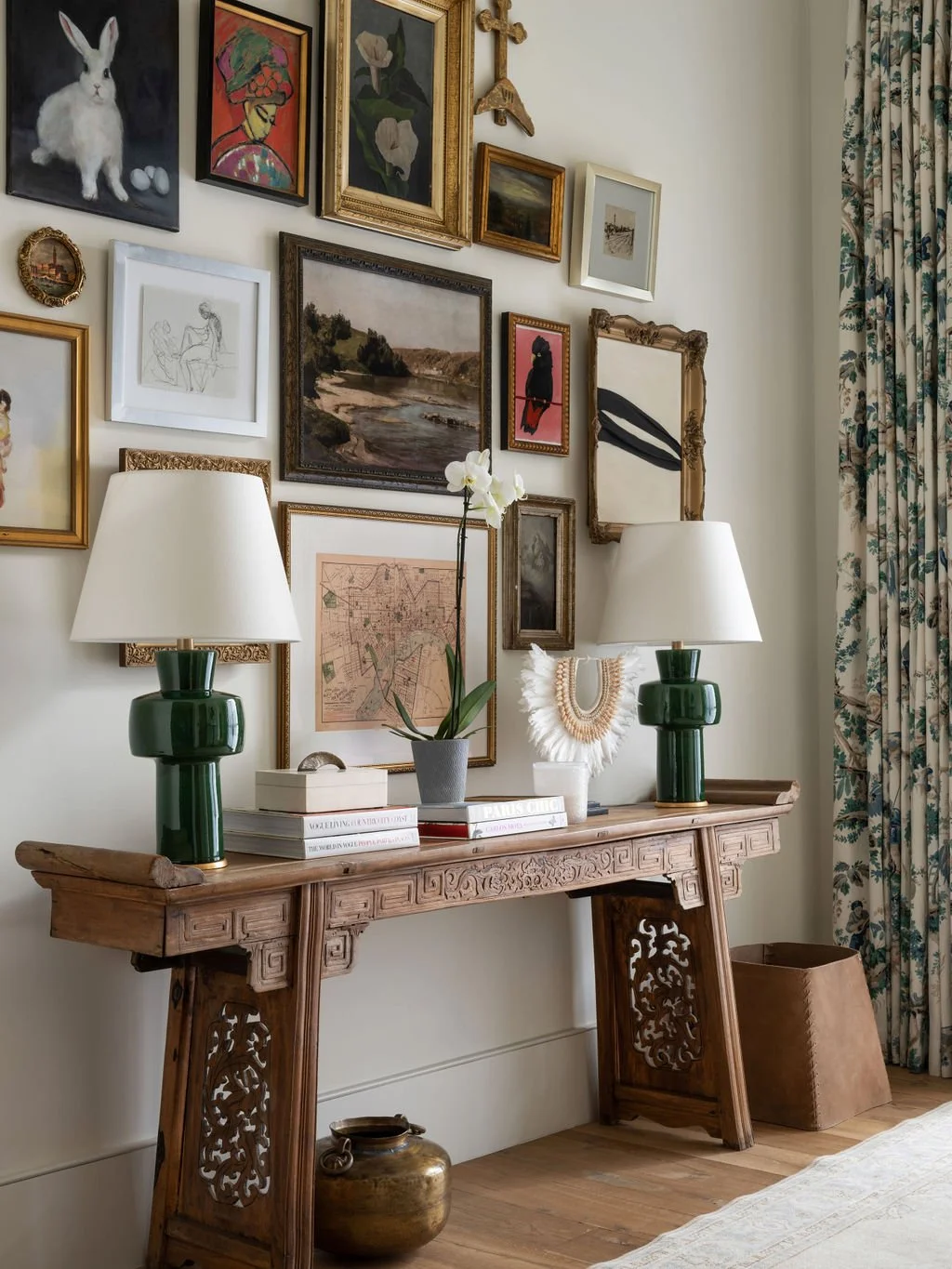 Decorative wall with various framed artwork above a wooden console table, which holds two green lamps, a potted orchid, books, and decorative items. Curtain with floral pattern on the right.