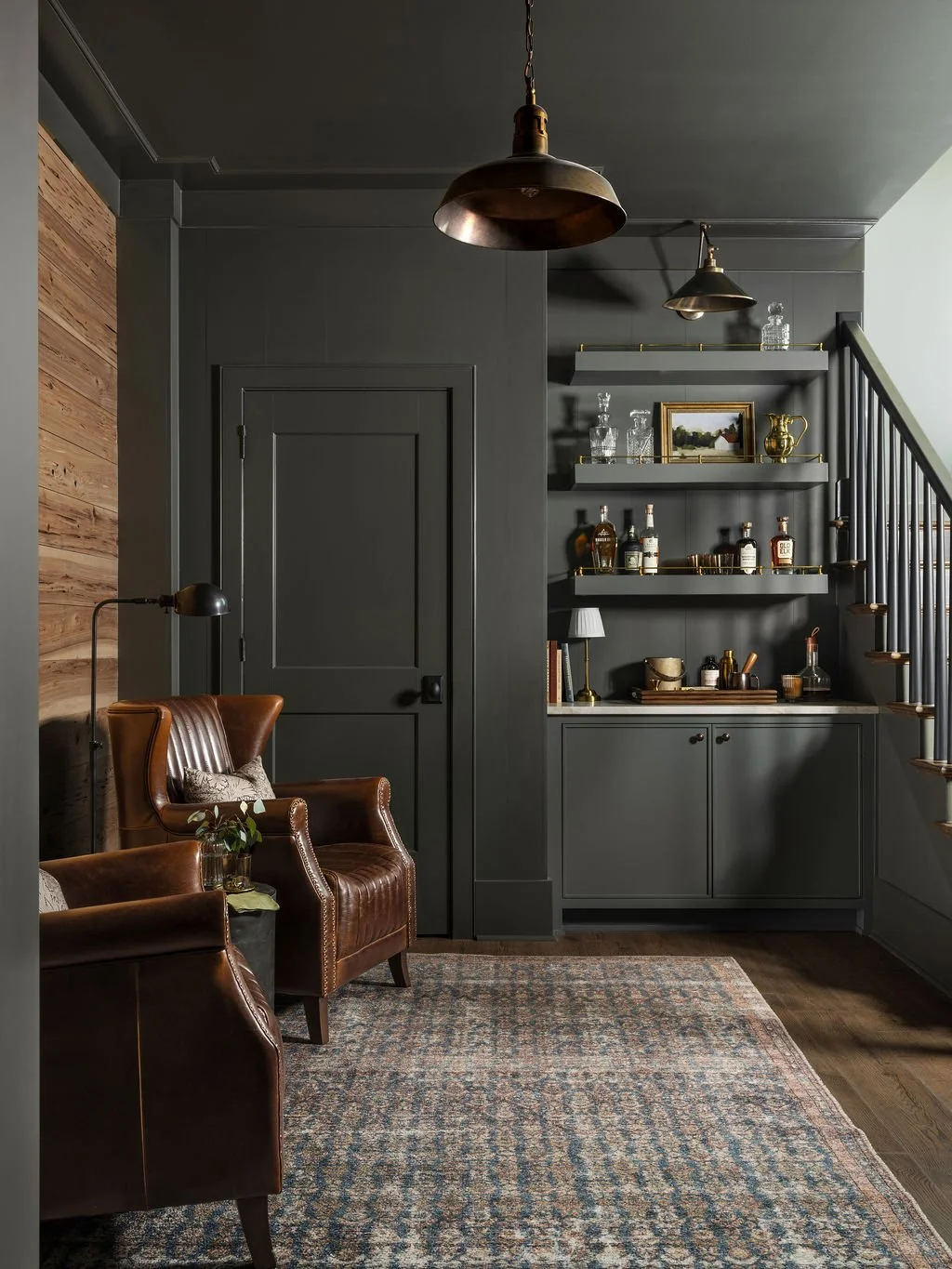 A cozy living room corner with dark gray walls, wooden accents, leather armchairs, a patterned rug, and a built-in shelving unit with decorative glassware, books, and ornaments. There are pendant lights hanging from the ceiling.