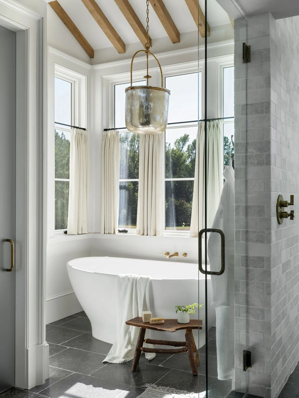 Bright bathroom with a white bathtub by bay windows, gold fixtures, and a pendant light. The space has white walls, a glass shower door, and a small wooden stool with toiletries and a plant.