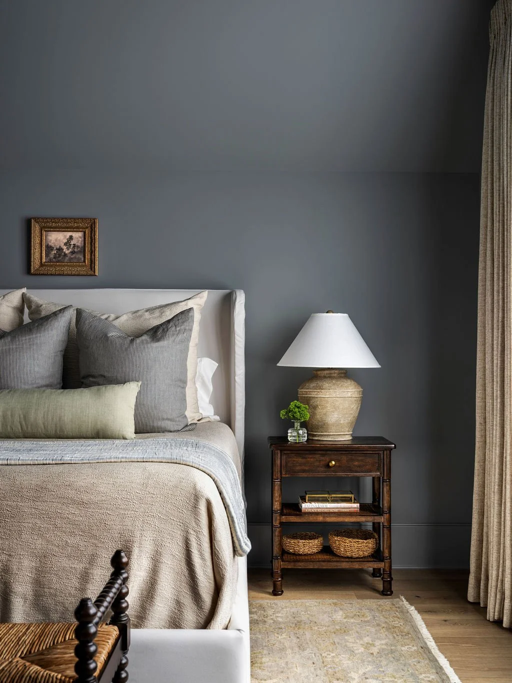 A bedroom with a white bed, gray pillows, a wooden nightstand with a ceramic lamp, a small plant in a glass jar, a bookshelf, and a beige curtain next to a window.