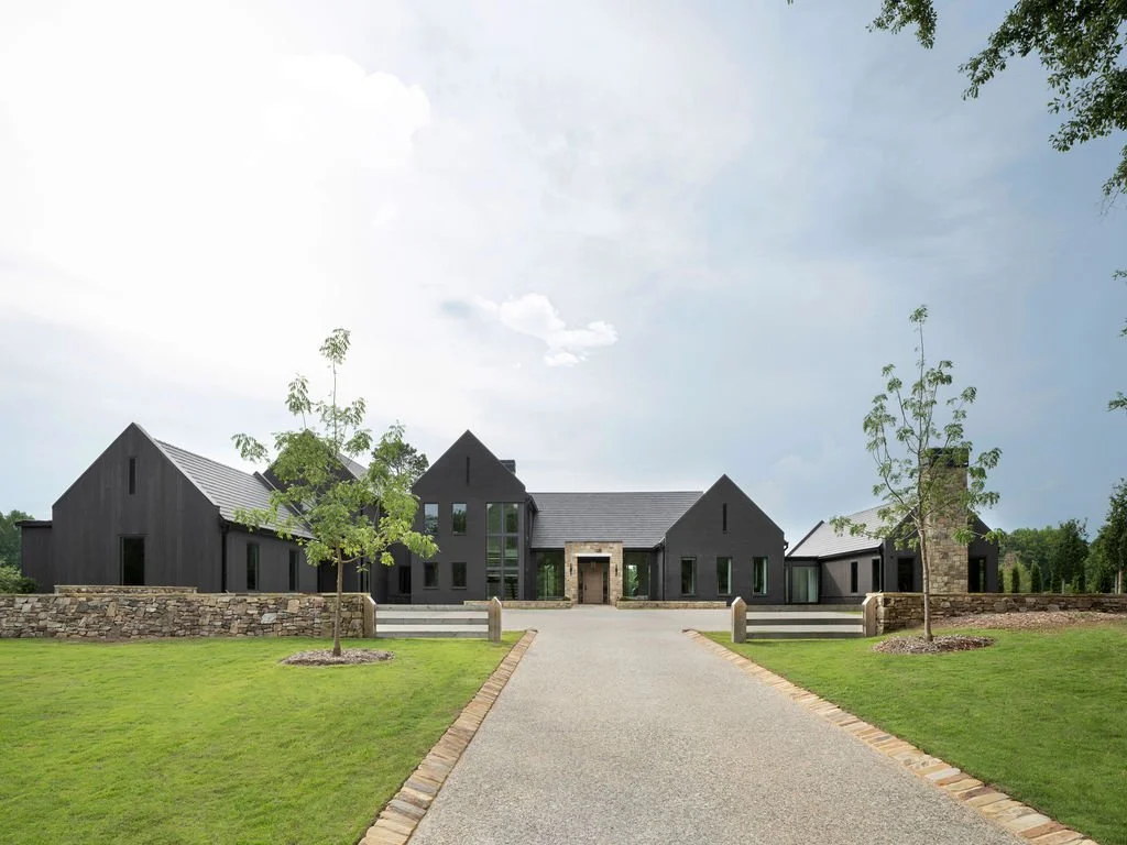 A modern black house with a stone entrance, surrounded by a manicured lawn, trees, and a stone pathway leading to the front steps.
