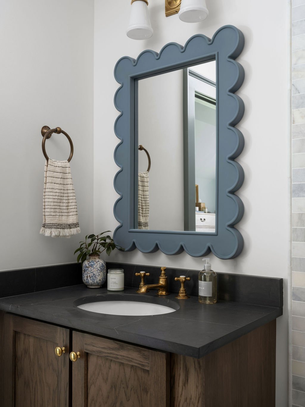 Bathroom sink with a dark countertop, a wooden vanity, brass faucet, a small potted plant, candles, and hand soap, with a decorative blue mirror and wall-mounted towel ring with a striped towel.