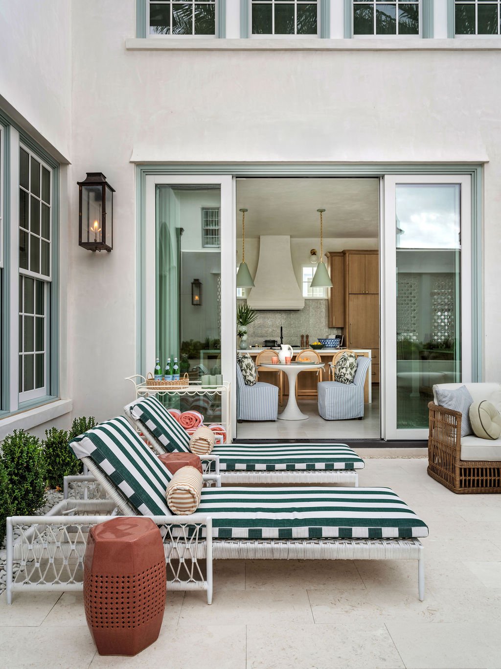 Outdoor patio with two striped lounge chairs, a small terracotta side table, and a white cushioned loveseat. Inside, a kitchen with a round table, striped chairs, pendant lighting, and wooden cabinets is visible through sliding glass doors.
