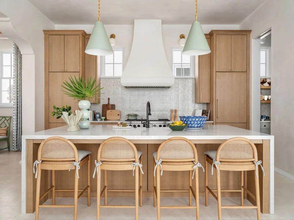 Modern kitchen with a white island, wooden cabinetry, four bar stools with tied ribbons, decorative plants, and pendant lights.