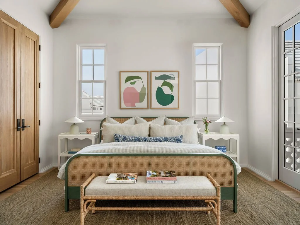 A bedroom with a white wall, two windows, and a wooden door. There is a bed with white pillows, a woven headboard, and a bench at the foot of the bed. Two white nightstands with lamps and small decor items are on either side of the bed. Artwork featu