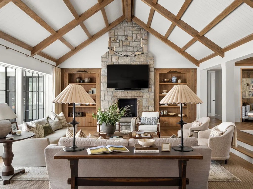 Living room with vaulted ceiling, stone fireplace with a mounted TV, beige sofas, armchairs, lamps, and built-in wooden shelves.