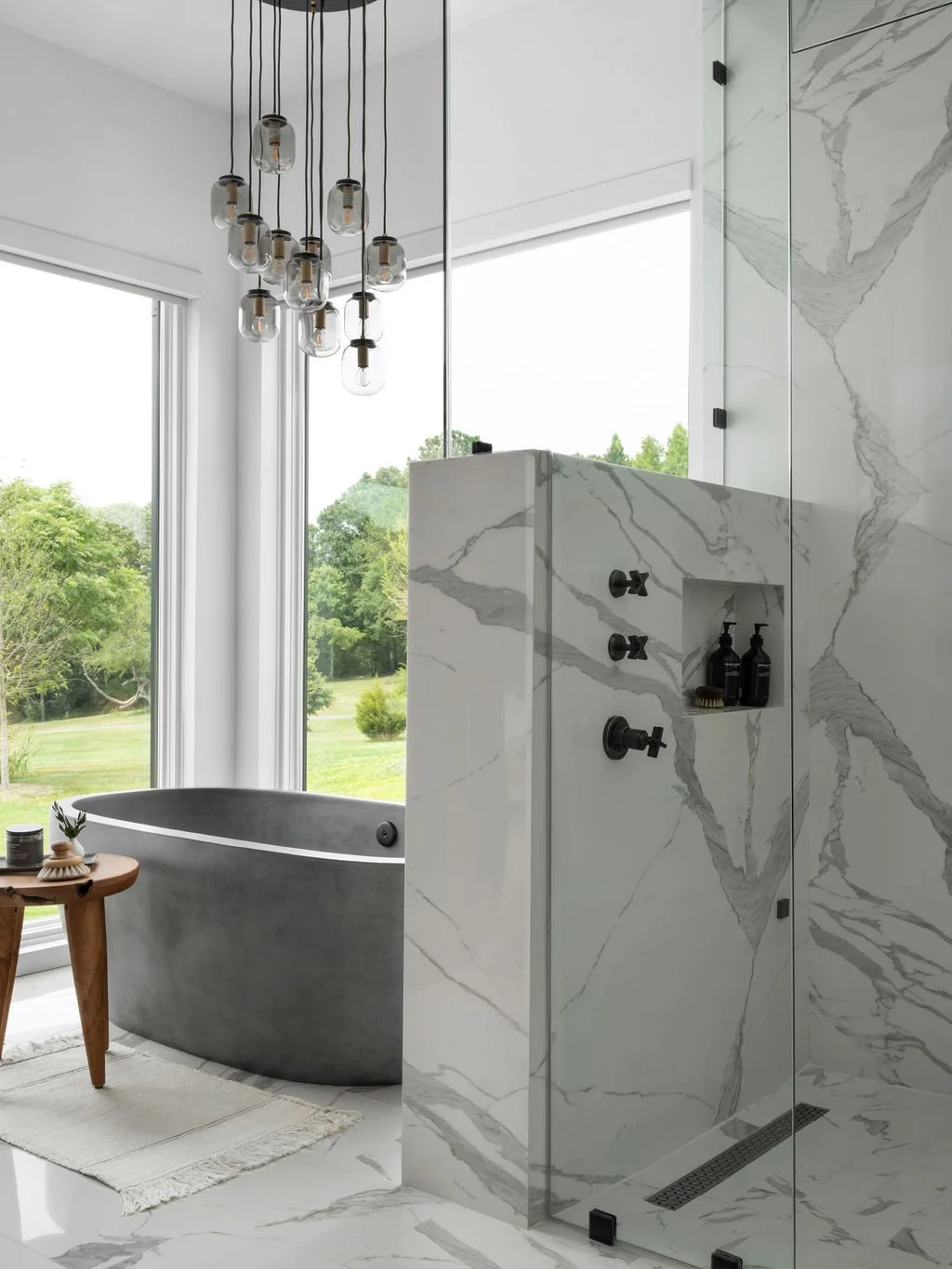 Modern bathroom with a gray freestanding bathtub, large windows with a view of green trees, marble walls, and a glass shower enclosure with black fixtures and bottles.