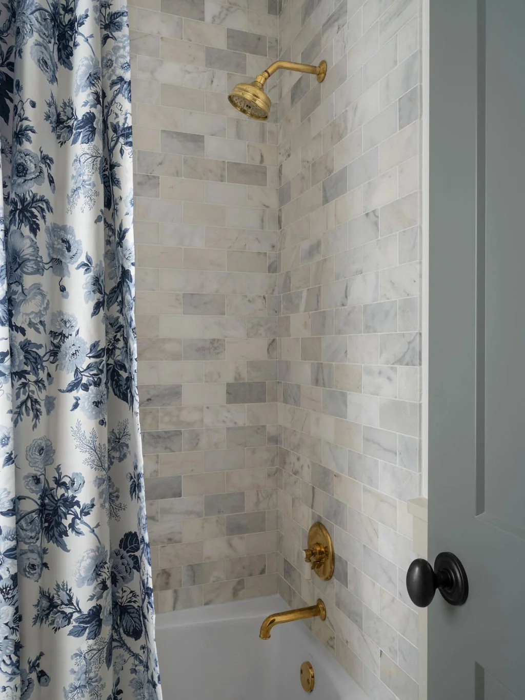Bathroom shower area with marble tile walls, brass fixtures, a brass showerhead, a brass tub spout, and a floral patterned shower curtain.