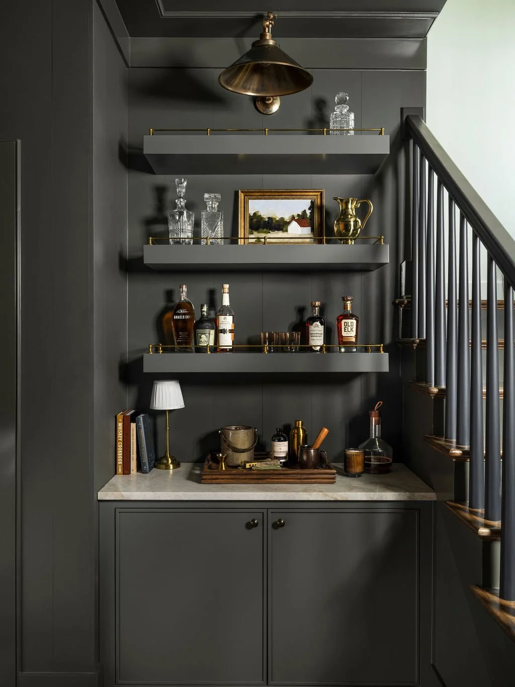 Dark gray wall with three floating shelves holding glass decanters and a gold pitcher, framed landscape painting, bottles of liquor, books, a small table lamp, and bar accessories beneath a brass light fixture, adjacent to staircase with dark railing