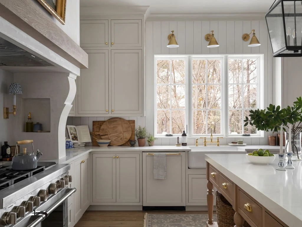 A bright kitchen with white cabinetry, a large window with a view of trees, and gold fixtures. There is a stove with a kettle, a white countertop with various items, and a wooden kitchen island with a bowl of limes and a plant.