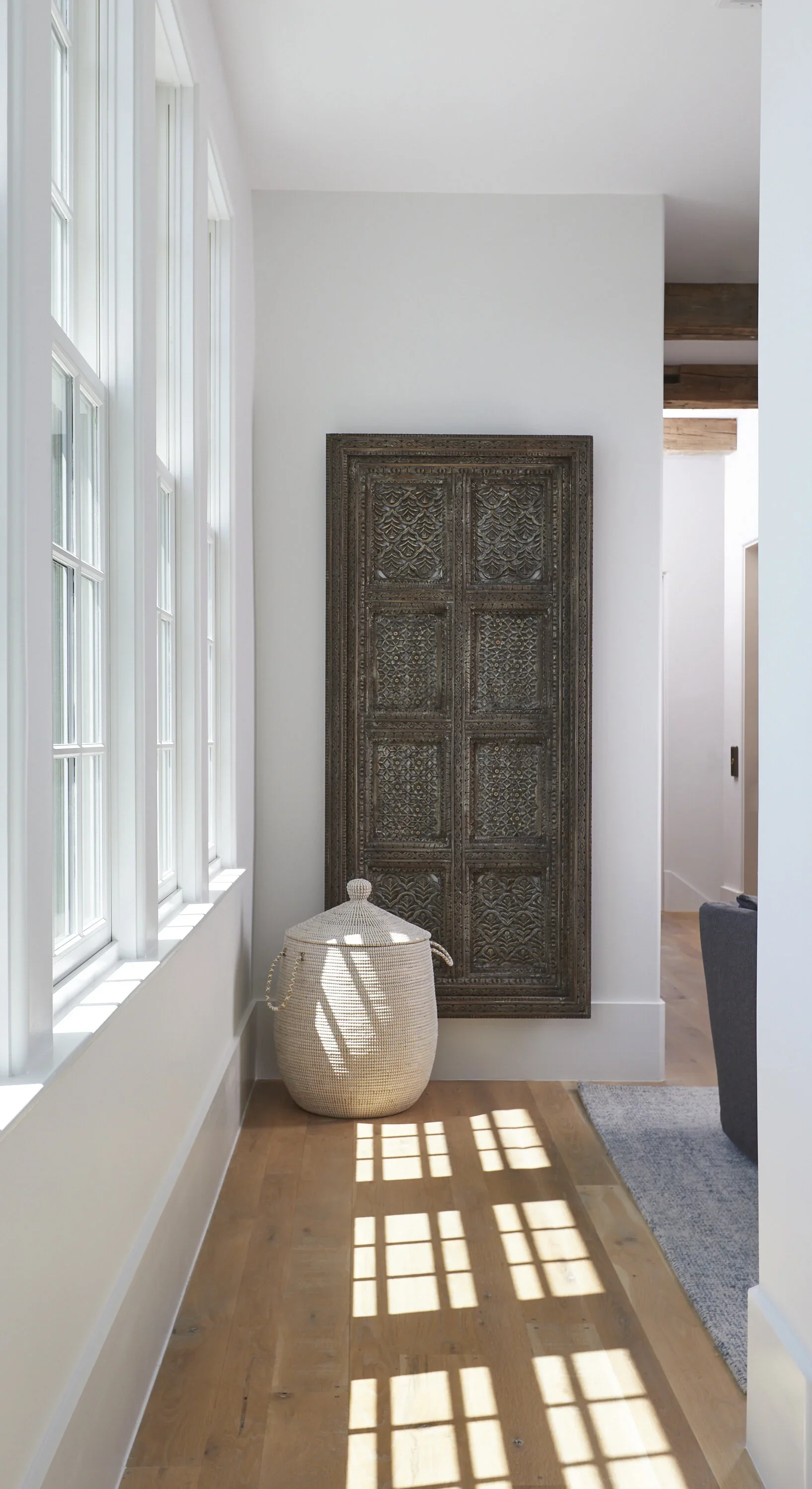 Sunlight streams through large windows, casting shadows onto a wooden floor in a minimalist interior with white walls, a decorative carved wooden panel, and a woven basket.