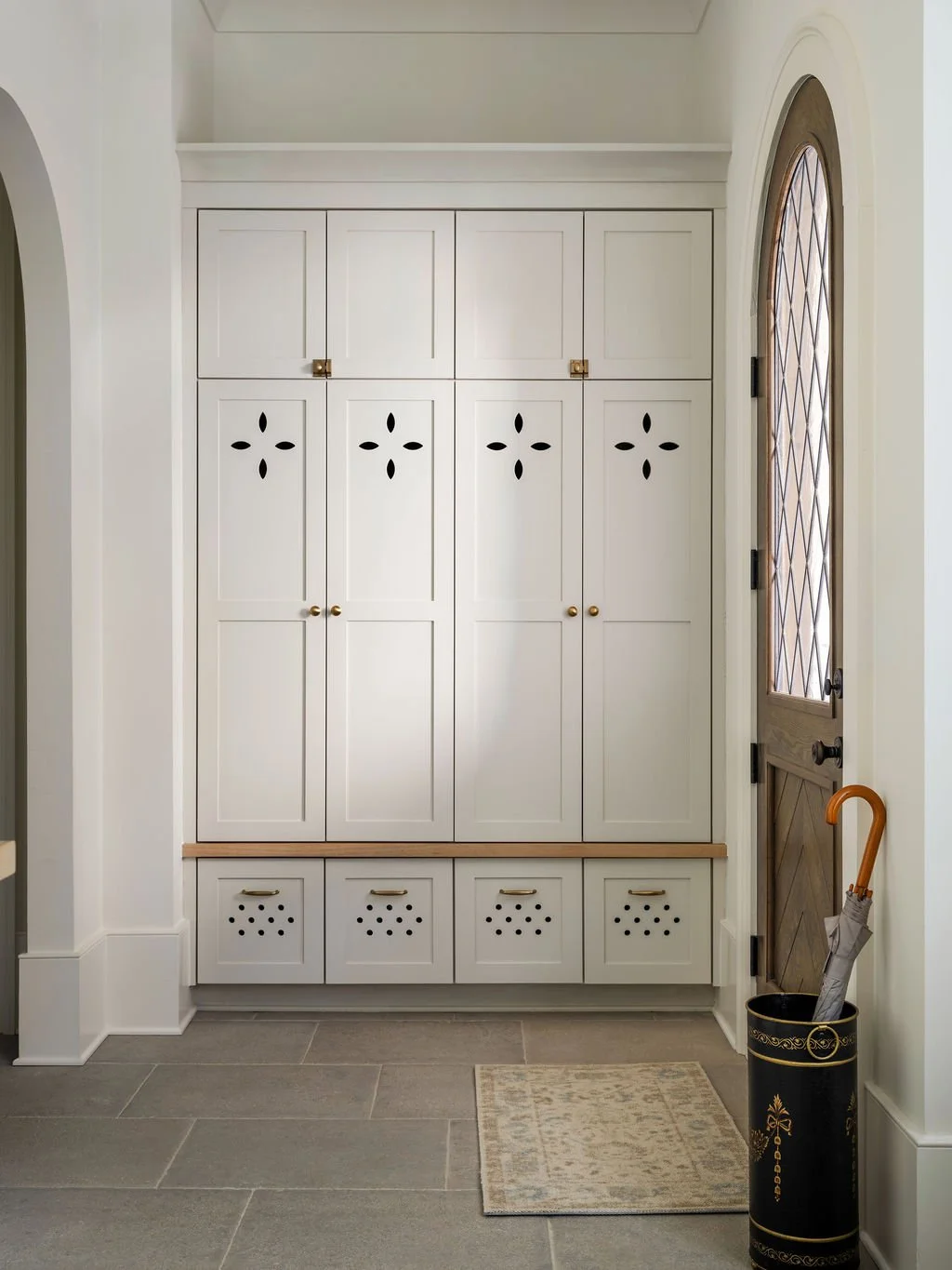 White built-in storage cabinets with decorative cutouts, brass knobs, and gold hardware on a tiled floor in an entryway next to a wood door with a diamond-patterned window, with an umbrella stand containing an umbrella in the foreground.