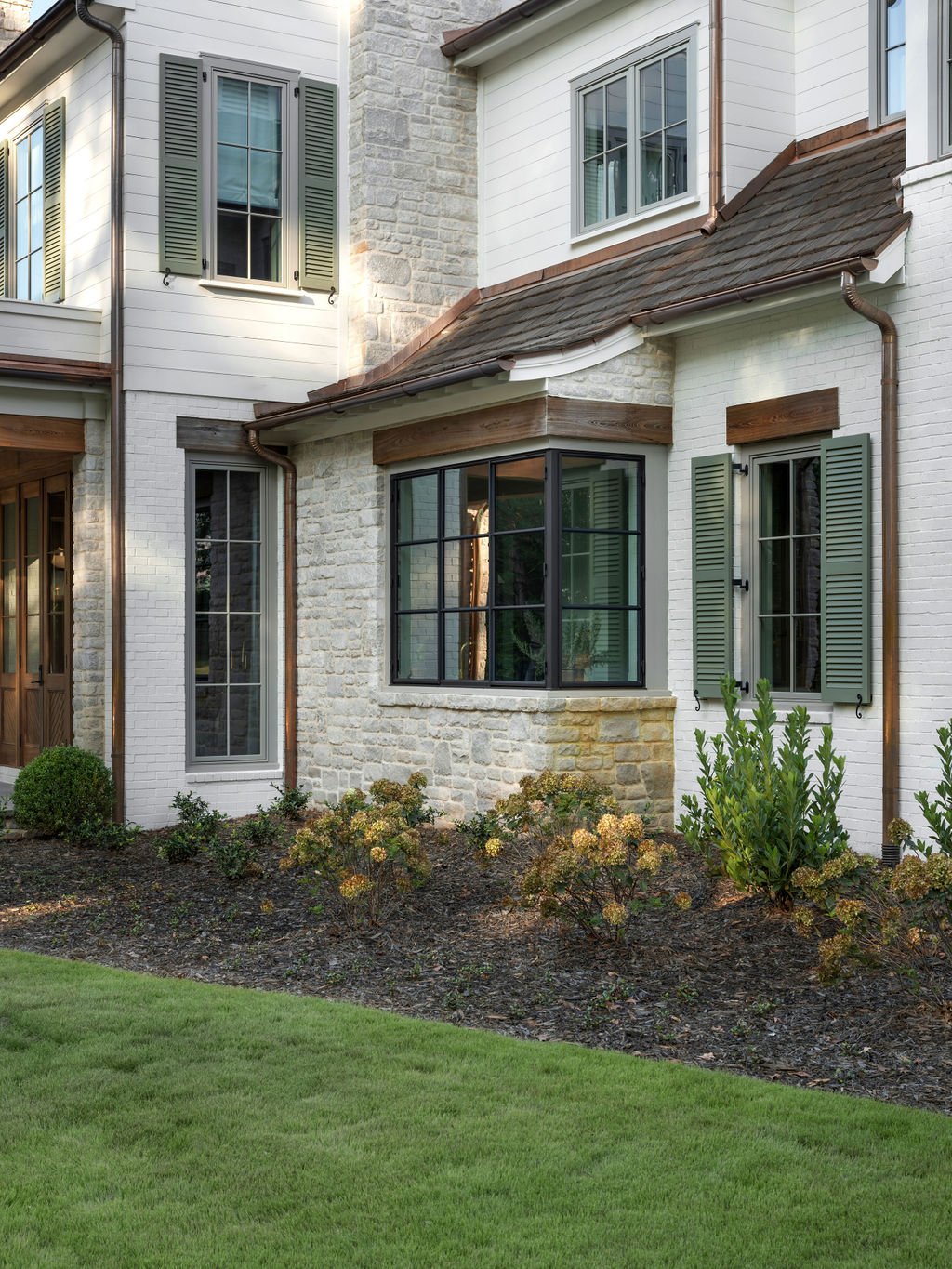 Exterior of a modern house with white brick and stone walls, green shutters on the windows, a large glass bay window, and a small landscaped garden with shrubs and grass.