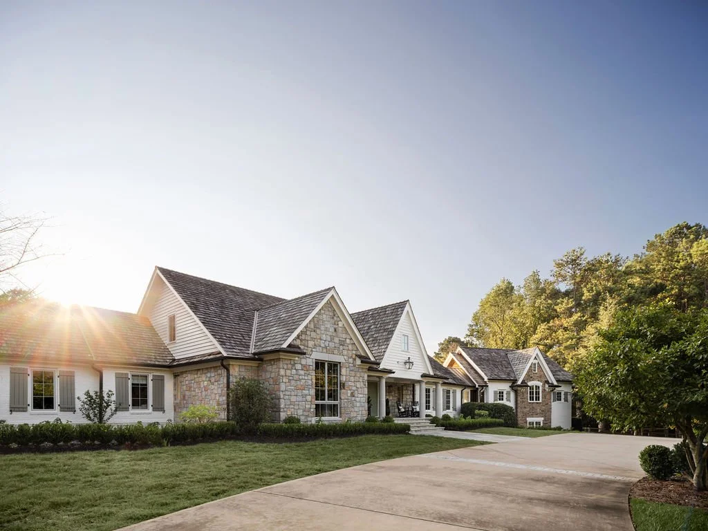 A large, elegant house with stone and white siding exterior, surrounded by well-maintained lawn and trees, with the sun setting in the background.