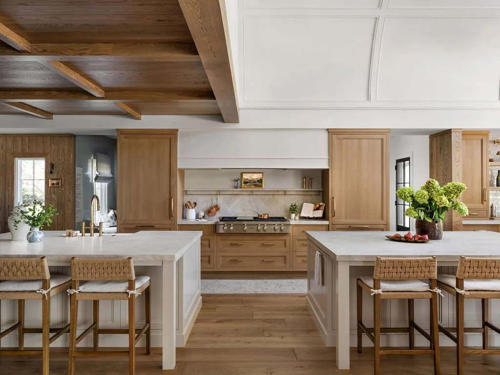 A modern kitchen with white countertops, wooden cabinets, and a large island with seating. The kitchen features open shelves, a stove, and decorative plants and vases.