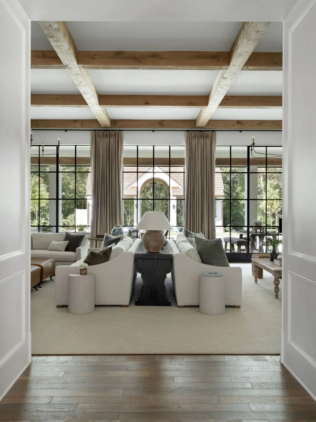 Living room with large windows, wooden ceiling beams, white sofas, and beige curtains.