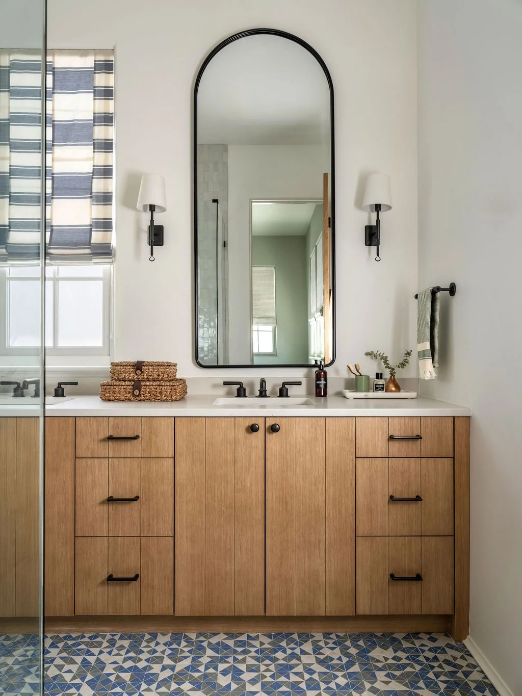 Bathroom vanity with wooden cabinet, white countertop, large mirror, black fixtures, wall sconces, and decorative items.