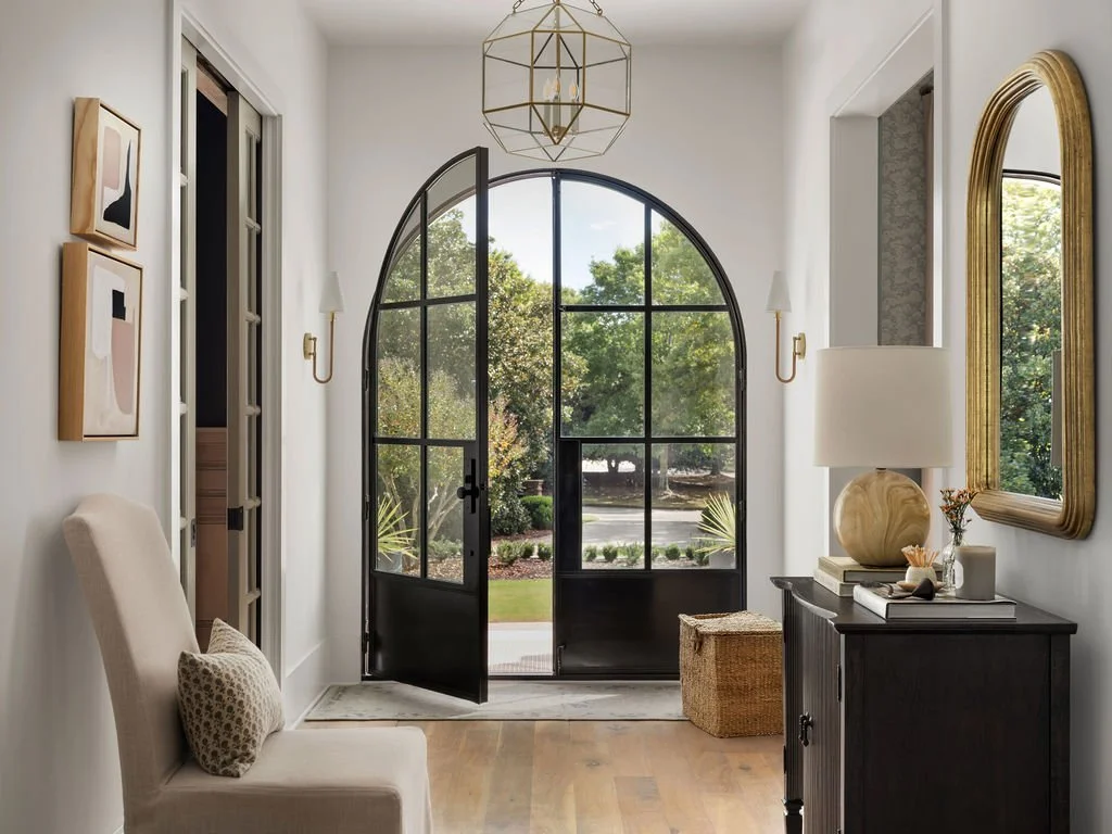 Bright entryway with black arched glass door, white walls, beige chair, gold mirror, table with lamp and decor, artwork, and view of greenery outside.