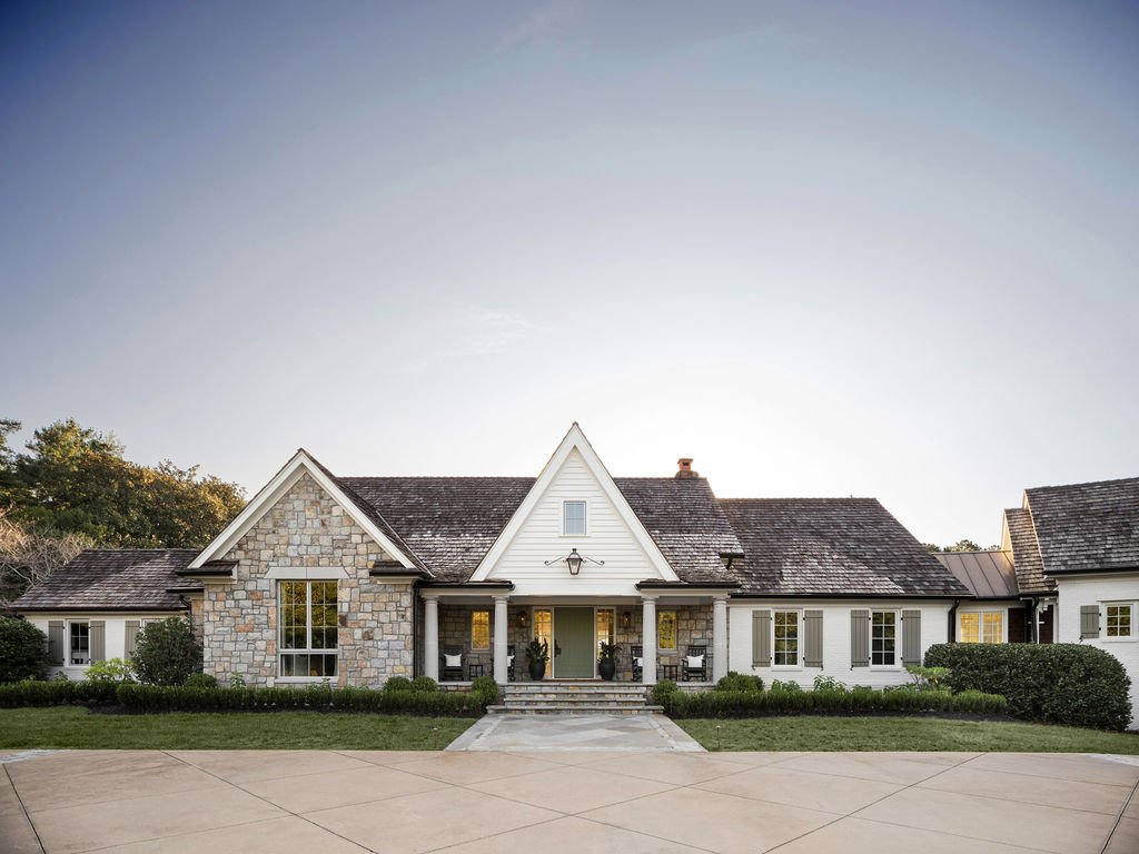 A large, modern house with a front porch, stone and white siding exterior, surrounded by a manicured lawn and bushes, under a clear sky.