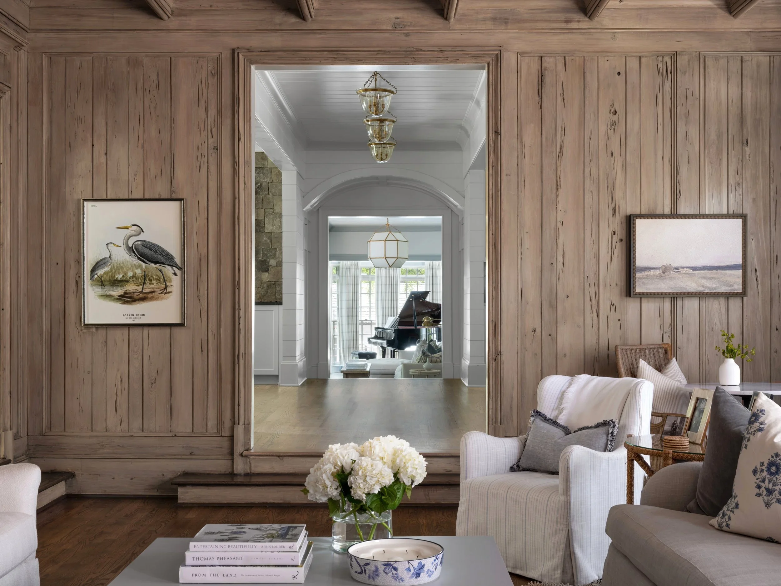 View of a living room with beige and white color scheme, wood-paneled wall with framed artwork, white sofa, glass coffee table with books and flowers, and a mirror reflecting another room with a grand piano and large windows.