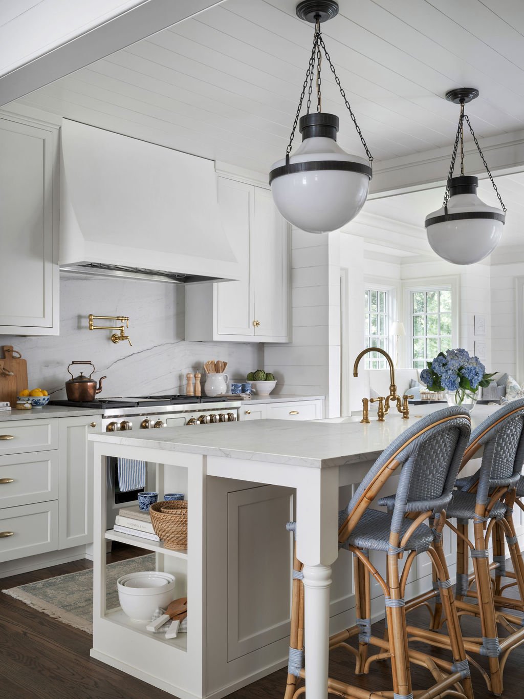 Cozy kitchen with white cabinetry, marble countertops, gold fixtures, and rattan chairs at a white island, lit by two hanging pendant lights.