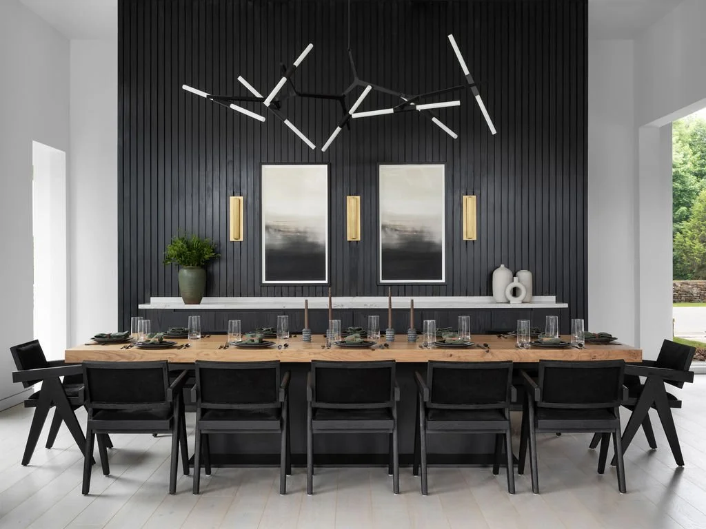 Modern dining room with a long wooden table set with black plates, glasses, and candles, black chairs, a black paneled accent wall, three abstract art pieces, a potted plant, and decorative vases.