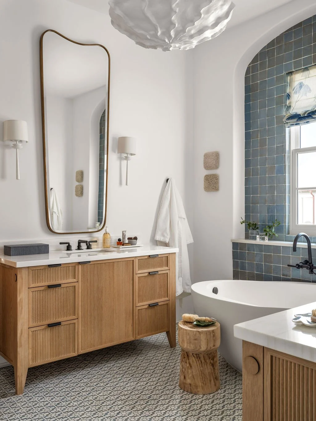 Modern bathroom with wooden vanity, white countertop, and black fixtures. Large mirror above sink, wall-mounted lights, freestanding bathtub, and patterned floor tiles. Blue tiled shower wall and window with plants.