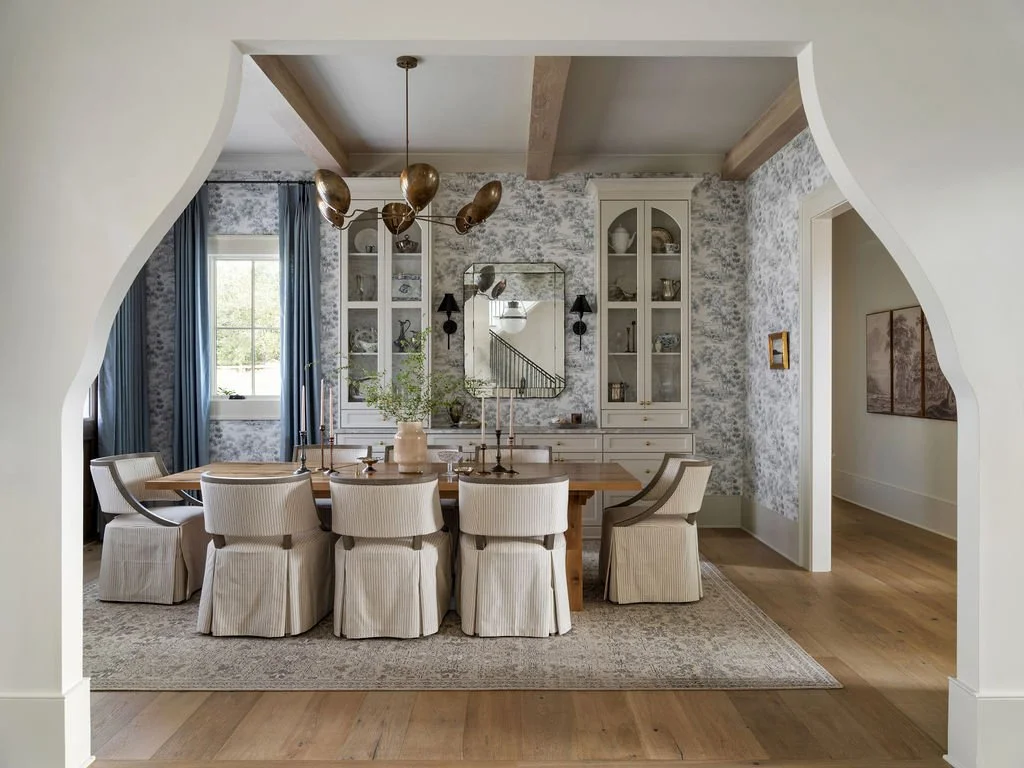 A dining room with a wooden table, white upholstered chairs, a large mirror, built-in white cabinets, and a window with blue curtains, decorated in a neutral and blue color theme.