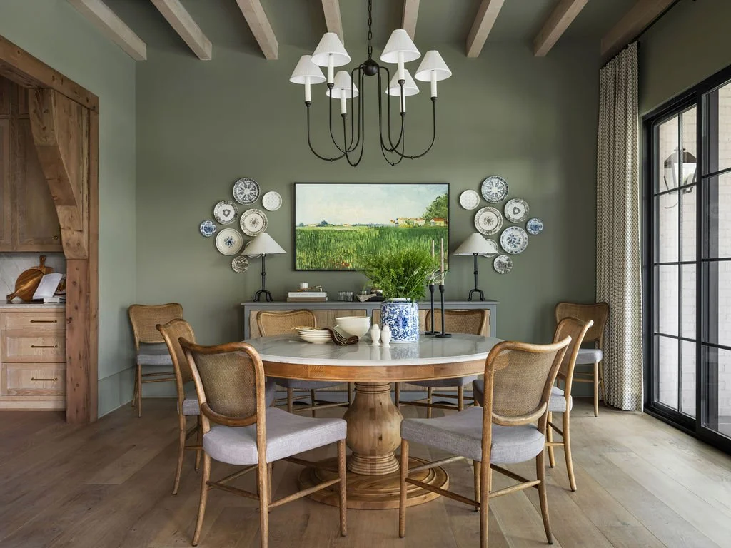 A dining room featuring a round wooden table with six chairs, a chandelier hanging above, wall decorations including plates and a painting, a sideboard with lamps, and large sliding glass doors with curtains.