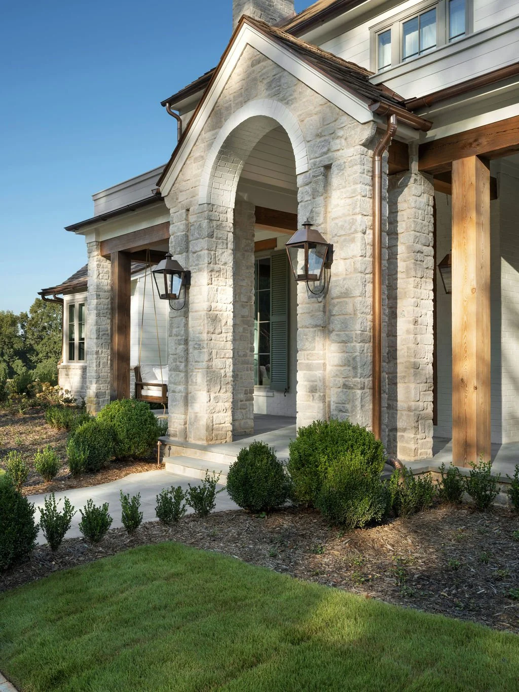 Front porch of a house featuring a stone archway, two lantern-style lights, hanging swing, and landscaped bushes, with a well-maintained lawn and a blue sky.