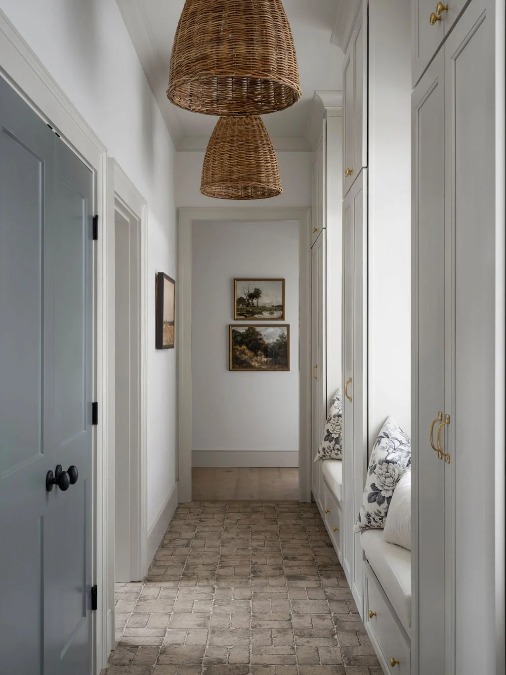 A hallway with white walls, cabinetry with gold handles, and a brick-patterned rug. Two wicker pendant lights hang from the ceiling, and there are two framed landscape paintings on the far wall, with built-in seating with floral pillows on the right.
