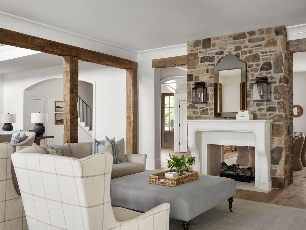 Living room with a stone fireplace, white mantel, and mirror, beige sofas, and wooden beams.