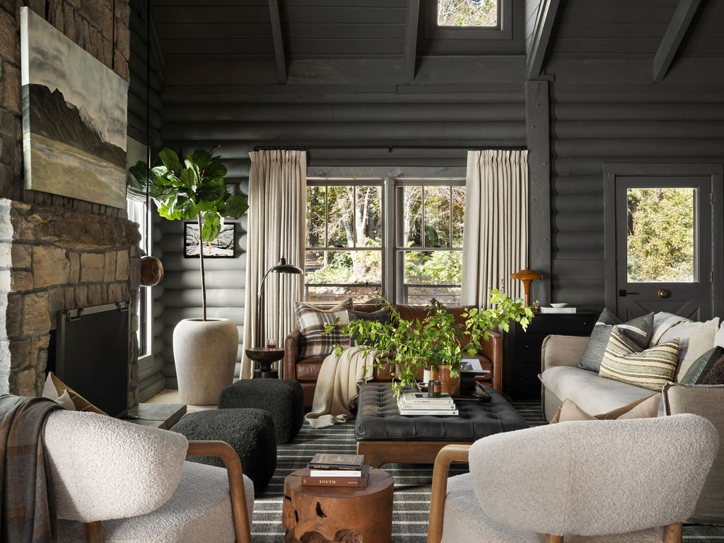 Living room with gray log walls, a stone fireplace, large windows, and a skylight. Contains various chairs, a leather couch, a black ottoman, and houseplants.