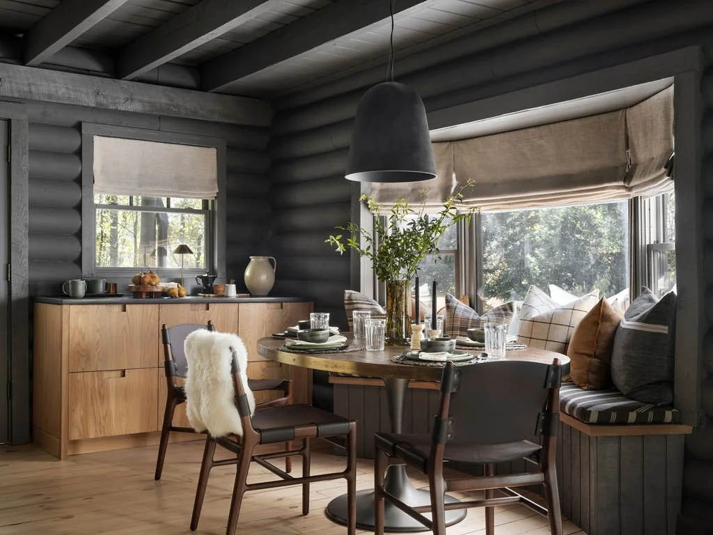 Cozy dining nook with a round wooden table, chairs, and a window seat with pillows inside a log cabin.
