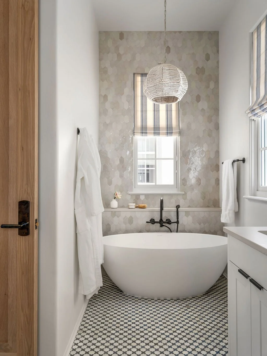 Bathroom with a white free-standing bathtub, black faucet, honeycomb-structured wall tiles, a window with striped window shades, a hanging light fixture, a white wall, and wooden door.