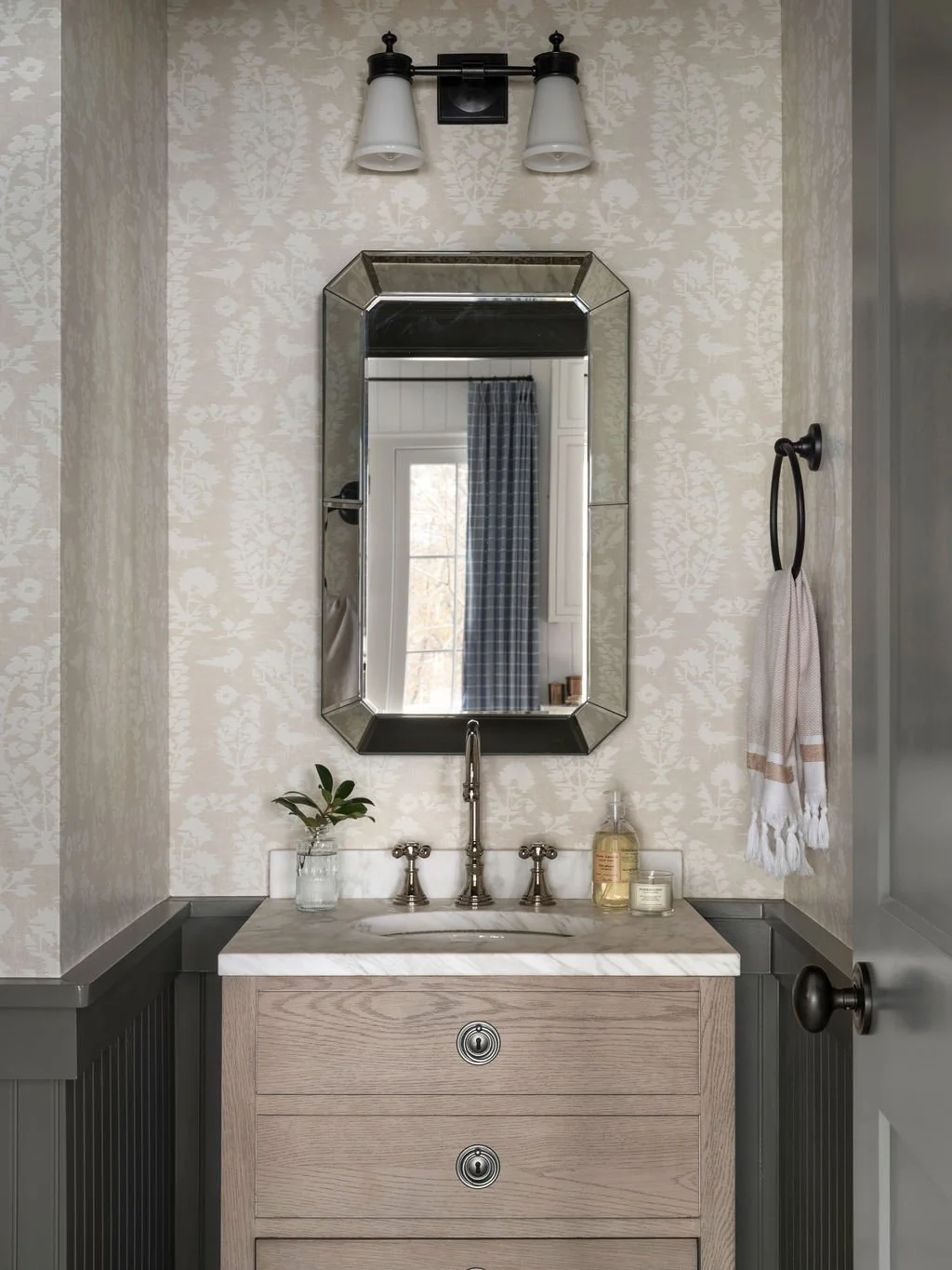 Bathroom vanity with a wooden cabinet, marble countertop, and a silver-framed mirror. There is a small potted plant, soap dispenser, and candle on the countertop. A wall-mounted light fixture with two lamps is above the mirror, and a towel ring with 