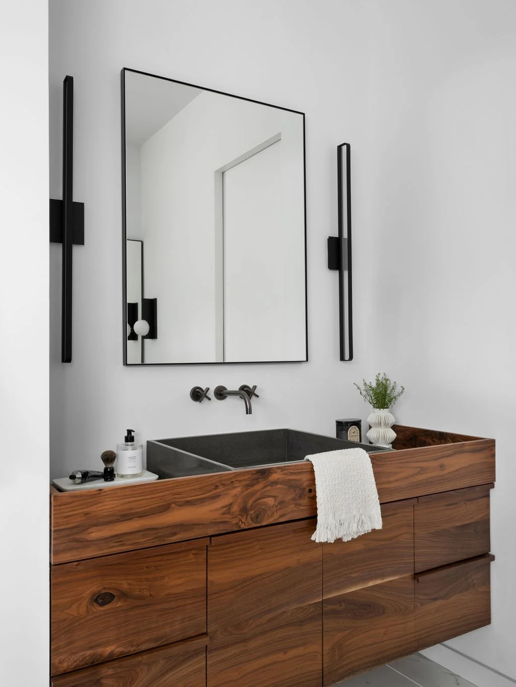 Modern bathroom vanity with a wooden cabinet, black sink, and wall-mounted faucet. Above the sink is a large mirror, flanked by black vertical light fixtures. On the vanity are toiletries, a small potted plant, and a folded white towel.