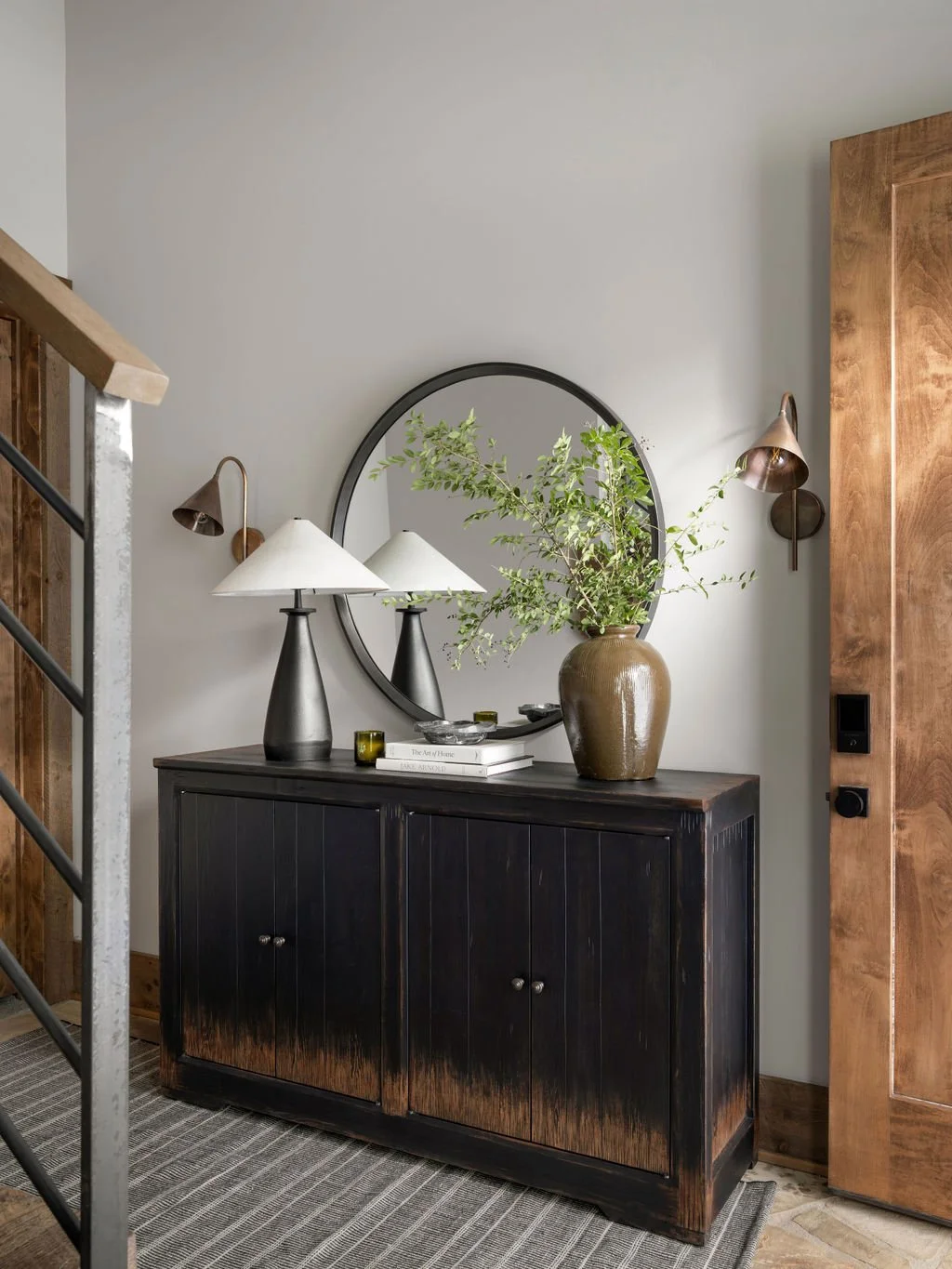 A black wooden sideboard with a large beige vase containing green foliage, a round mirror, two black lamps with white shades, a stack of books, and small decorative objects on top, against a light-colored wall with wooden accents.