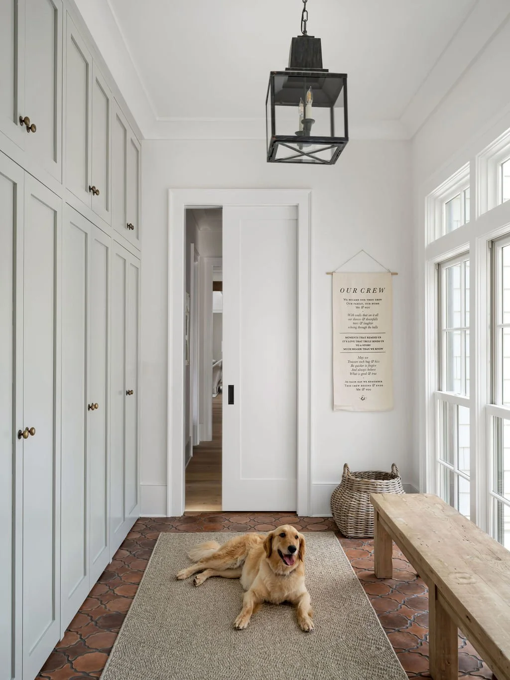 A hallway with white walls and cabinetry, a wooden bench, a woven basket, and a golden retriever lying on a beige rug. Sunlight streams through large windows on the right, and a black lantern-style ceiling light hangs overhead. A wall hanging with te