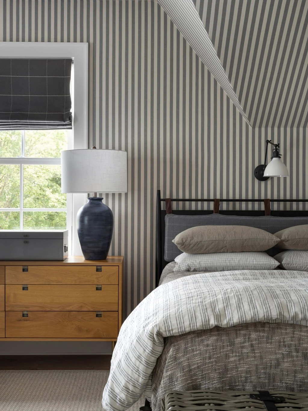 A bedroom with a wood dresser, a lamp, a window with a black Roman shade, striped wallpaper, a bed with pillows, and a reading lamp on the wall.