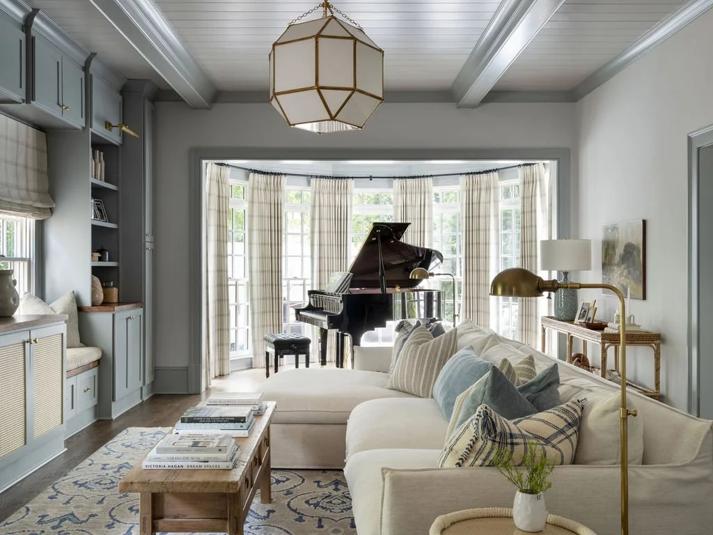 Bright living room with a grand piano near large bay windows, cream sofa with assorted pillows, a wooden coffee table with books, a blue built-in shelf, and a white ceiling with a geometric pendant light.