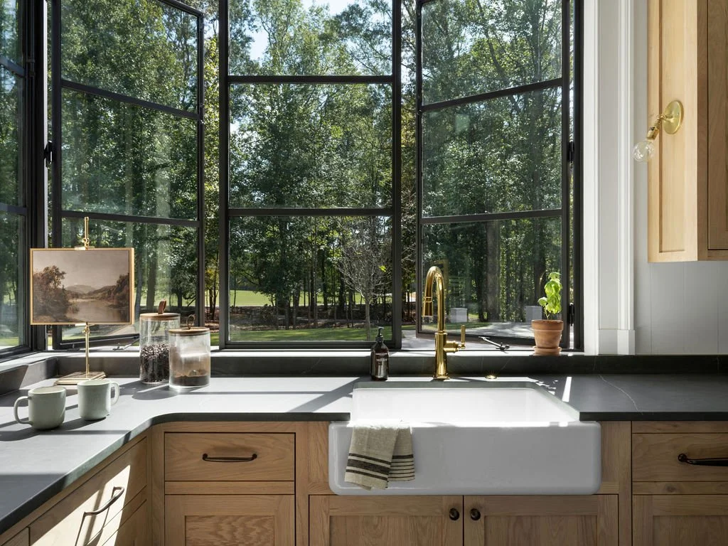 Kitchen sink with a large window above, overlooking trees outside. Wooden cabinets, a small potted plant, and decorative jars on the countertop.