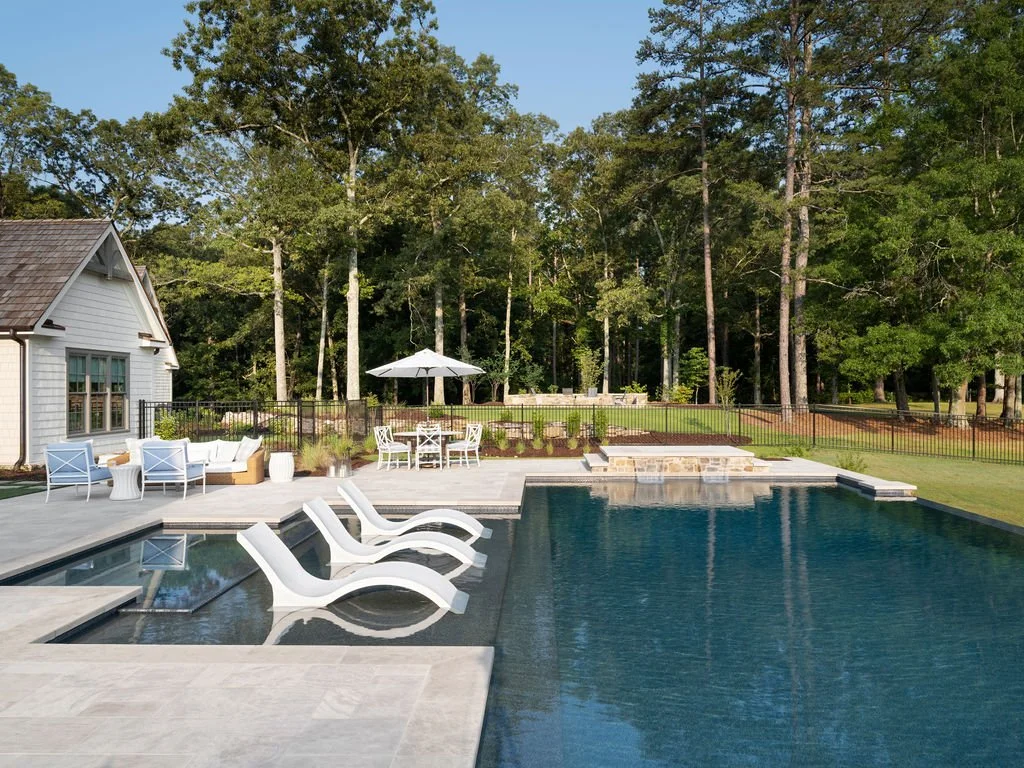 A backyard with a swimming pool, white lounge chairs, and an outdoor seating area with a umbrella. There are trees and a house in the background.