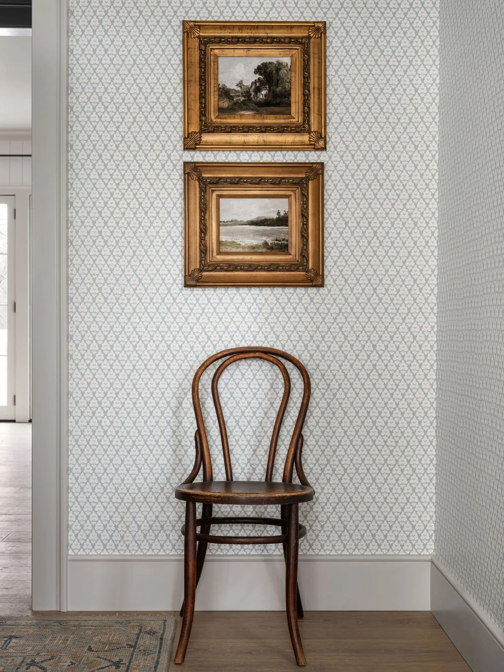 A wooden chair with a rounded backrest, placed against a wall with patterned wallpaper, beneath two framed landscape paintings. The paintings depict natural scenery with trees and water, and are adorned with ornate gold frames.