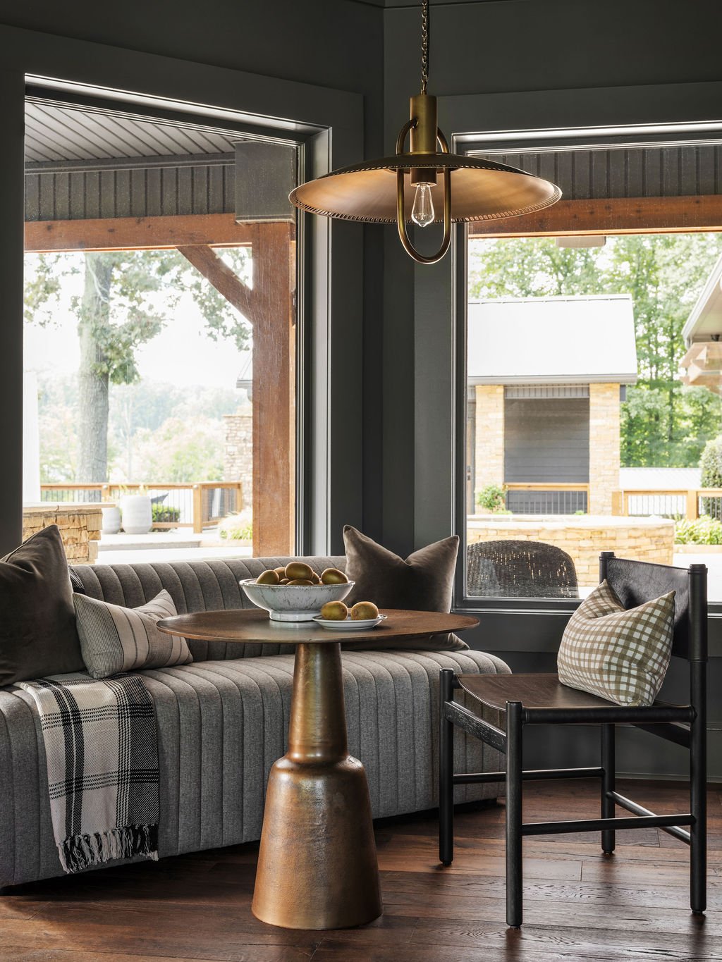 Living room with large windows, a gray sofa with pillows, a wooden side table with a bowl of lemons, a black chair with a checkered pillow, and a hanging brass light fixture.