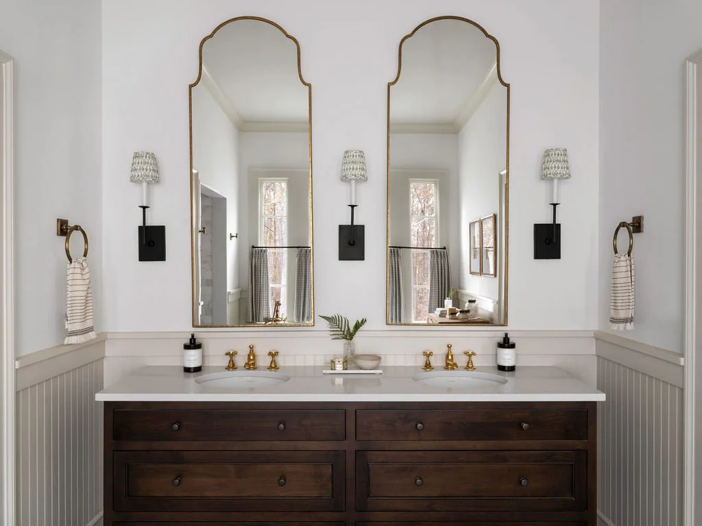 Double bathroom vanity with dark wood cabinets, white countertop, and two large mirrors with gold frames. There are two wall-mounted lamps with patterned shades, towel rings with striped towels, and black soap dispensers. A small plant and decor item